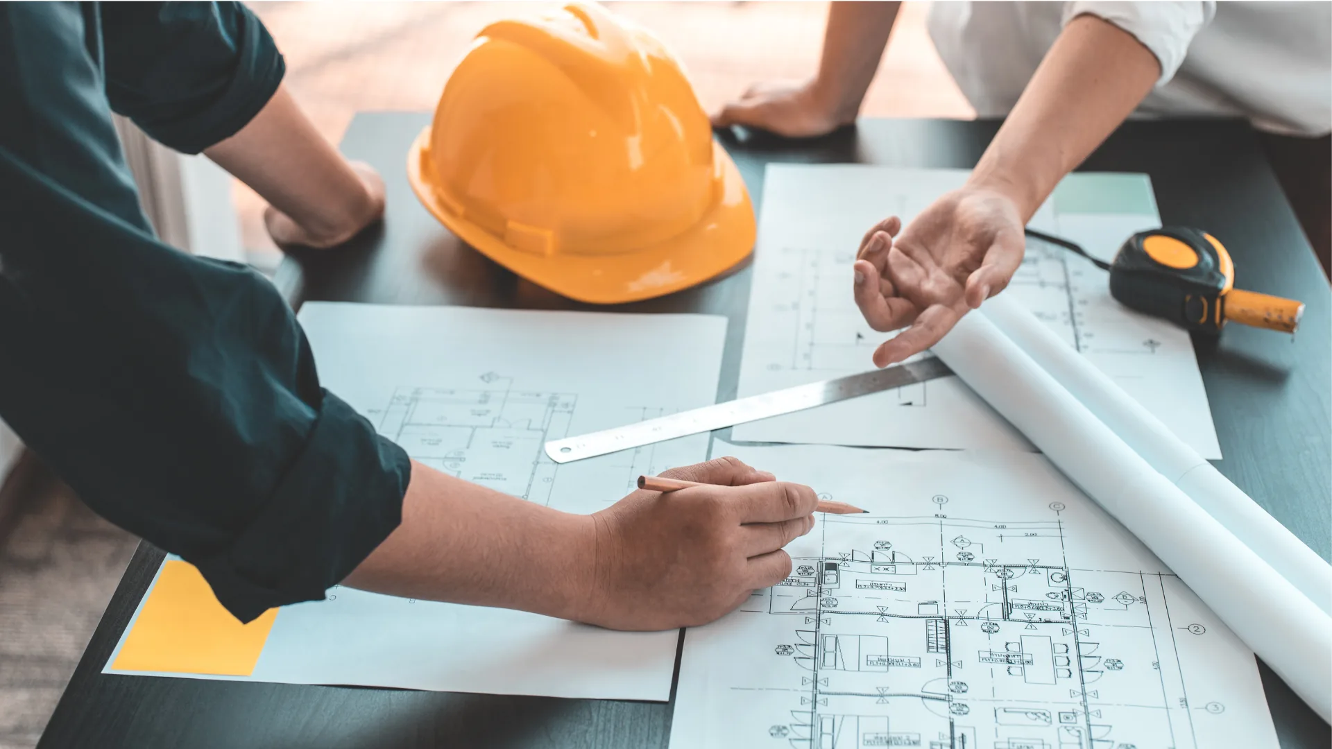 Profissionais analisando plantas técnicas sobre uma mesa, com capacete de obra ao fundo